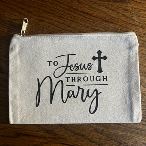 May include: A beige canvas zippered pouch with the text "TO Jesus THROUGH Mary" in black script and a cross symbol. The pouch has a gold-colored zipper.
