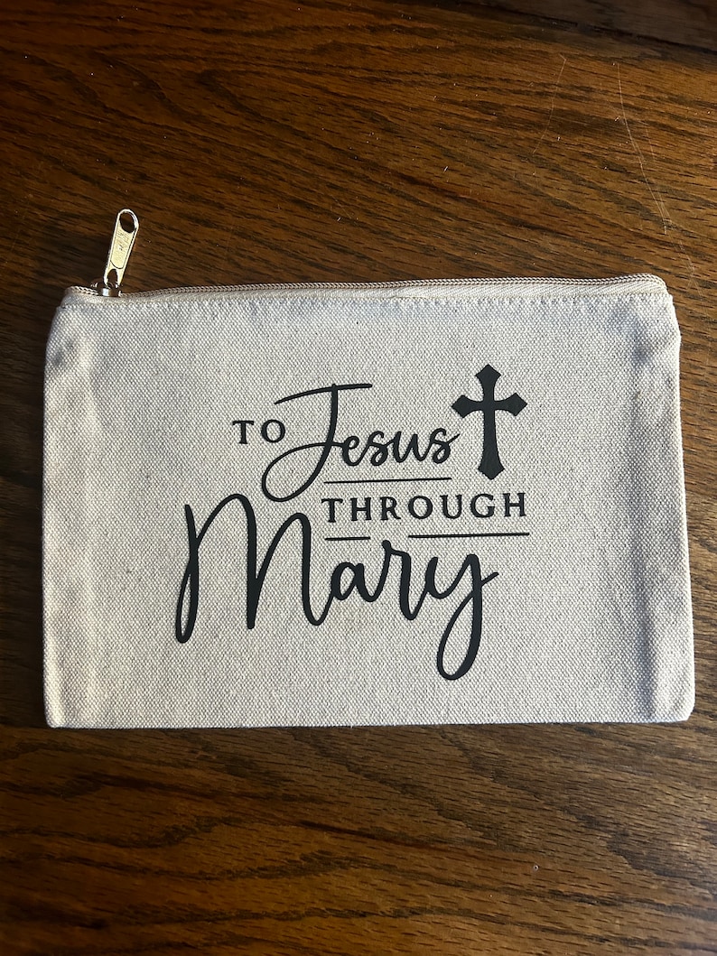 May include: A beige canvas zippered pouch with the text "TO Jesus THROUGH Mary" in black script and a cross symbol. The pouch has a gold-colored zipper.