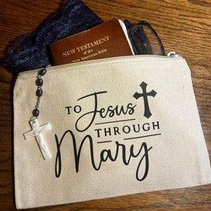 May include: A beige canvas zippered pouch with the text "TO Jesus THROUGH Mary" and a cross graphic. The pouch contains a New Testament of the New American Bible, glasses, and a rosary with a white crucifix.
