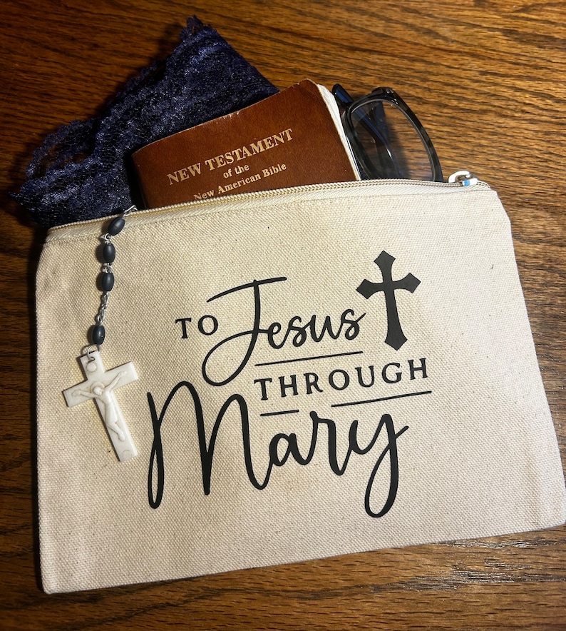 May include: A beige canvas zippered pouch with the text "TO Jesus THROUGH Mary" and a cross graphic. The pouch contains a New Testament of the New American Bible, glasses, and a rosary with a white crucifix.