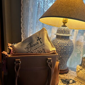 May include: A brown handbag with a zippered pocket and shoulder strap, holding a canvas pouch with the text "To Jesus Through Mary" and a cross. A blue and white decorative lamp and a statue are in the background.