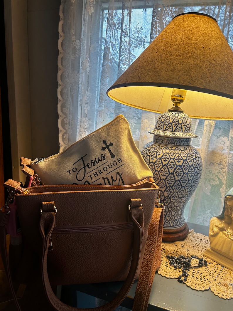 May include: A brown handbag with a zippered pocket and shoulder strap, holding a canvas pouch with the text "To Jesus Through Mary" and a cross. A blue and white decorative lamp and a statue are in the background.