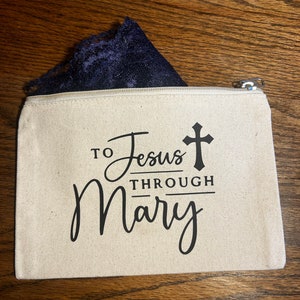 May include: A beige canvas zippered pouch with the text "TO Jesus THROUGH Mary" in black script and a black cross. A piece of navy blue lace is visible inside the pouch. The pouch is on a wooden surface.