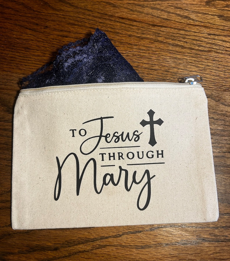 May include: A beige canvas zippered pouch with the text "TO Jesus THROUGH Mary" in black script and a black cross. A piece of navy blue lace is visible inside the pouch. The pouch is on a wooden surface.