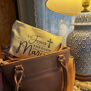 May include: A light brown handbag with a matching zippered pouch inside. The pouch is beige with the text "TO Jesus THROUGH Mary" in black script. A blue and white decorative lamp is in the background.