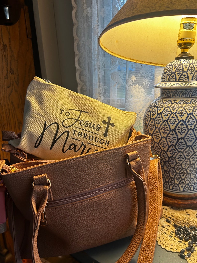 May include: A light brown handbag with a matching zippered pouch inside. The pouch is beige with the text "TO Jesus THROUGH Mary" in black script. A blue and white decorative lamp is in the background.