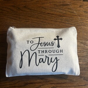 May include: A rectangular, off-white canvas zippered pouch with the text "To Jesus Through Mary" in black script and a cross symbol. The pouch is on a wooden surface.