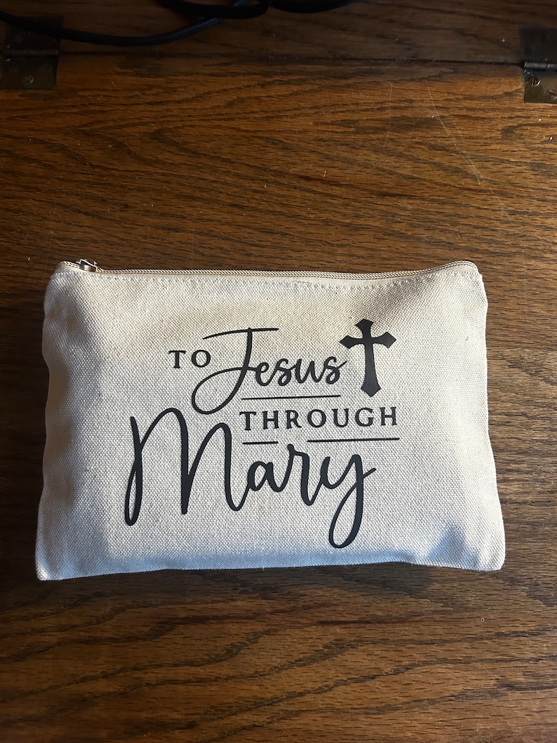 May include: A rectangular, off-white canvas zippered pouch with the text "To Jesus Through Mary" in black script and a cross symbol. The pouch is on a wooden surface.