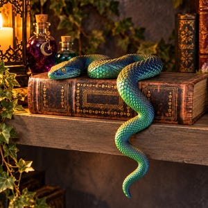 May include: A realistic snake figurine with iridescent blue and green scales resting on top of a thick, vintage-style leather-bound book. The snake has a textured, serpentine body that curves across the cover and hangs over the edge.