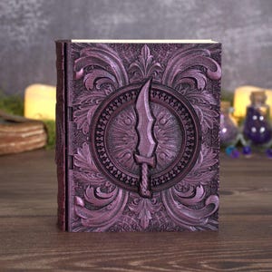 May include: A rectangular book with a deep purple cover featuring an embossed, intricate floral pattern and a central circular medallion containing a raised, stylized sword design. The spine is visible on the left side, and the pages are cream-colored.