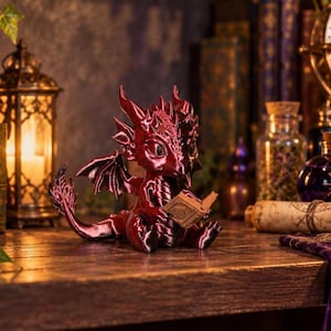 May include: A small, metallic dark red dragon figurine with detailed scales, horns, and wings, sitting in a seated position while holding a miniature open book between its front claws. The dragon has large, dark eyes and a textured, glossy finish.