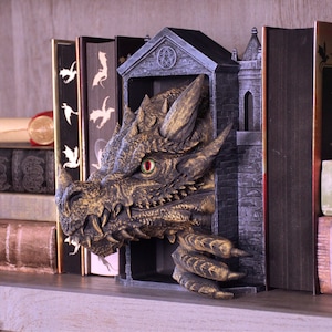 May include: A dark gray and gold-toned dragon bookend featuring a detailed dragon head emerging from a stone-like archway structure. The dragon has sharp scales, prominent horns, and a single visible eye with a red iris and black pupil.