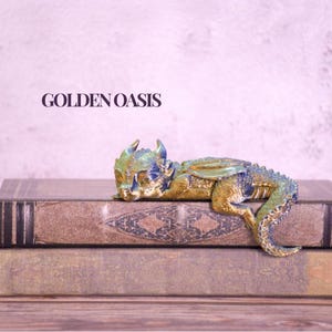 May include: A detailed figurine of a small dragon, in shades of gold, blue, and green, rests on two antique books. The dragon is posed as if sleeping. The text "GOLDEN OASIS" is visible in the background.