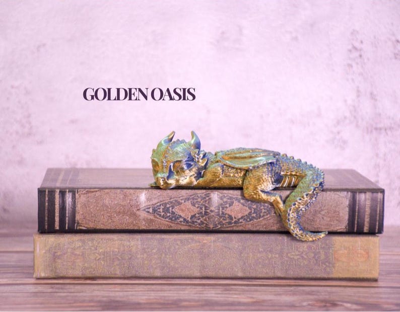May include: A detailed figurine of a small dragon, in shades of gold, blue, and green, rests on two antique books. The dragon is posed as if sleeping. The text "GOLDEN OASIS" is visible in the background.