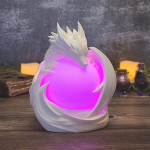 May include: A white, sculpted dragon figurine wrapping its body around a glowing, spherical light source that emits a vibrant, bright pink illumination. The dragon features sharp, angular scales and a pointed head, creating a protective posture.