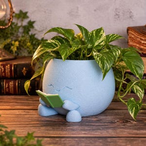 May include: A light blue, spherical planter with a textured surface, featuring a small character with closed eyes holding a green book. A variegated pothos plant with green and white leaves grows out of the top of the round, seated container.