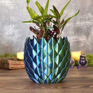 May include: A plant pot featuring a metallic, iridescent finish that shifts from deep blue to vibrant green. The exterior is composed of overlapping, scale-like segments arranged in a repeating pattern, housing a leafy plant with variegated foliage.