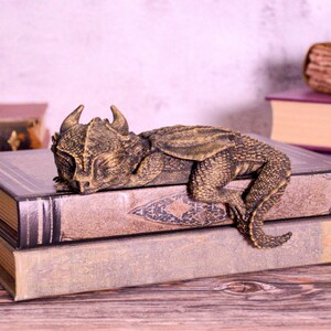 May include: A bronze-colored dragon figurine resting on a stack of vintage books. The dragon has detailed scales and horns. The books have aged covers and decorative patterns, creating a fantasy theme.