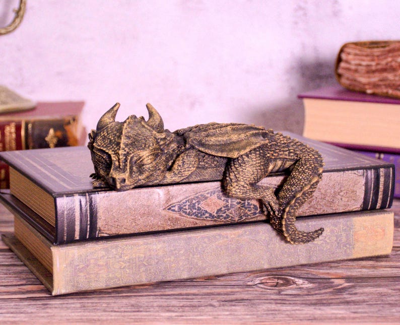 May include: A bronze-colored dragon figurine resting on a stack of vintage books. The dragon has detailed scales and horns. The books have aged covers and decorative patterns, creating a fantasy theme.