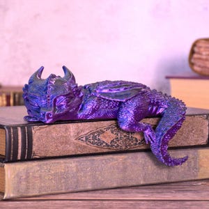 May include: A detailed, purple and blue dragon figurine resting on top of two antique books. The dragon has horns, wings, and a long tail. The books have aged, textured covers. The overall theme is fantasy and collectibles.