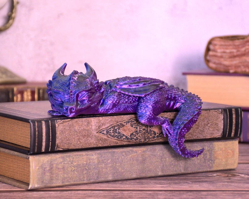 May include: A detailed, purple and blue dragon figurine resting on top of two antique books. The dragon has horns, wings, and a long tail. The books have aged, textured covers. The overall theme is fantasy and collectibles.
