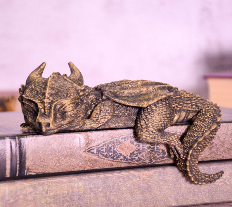May include: A detailed, bronze-colored dragon figurine resting on a stack of books. The dragon has horns, textured scales, and folded wings. The books have decorative covers with a vintage aesthetic.