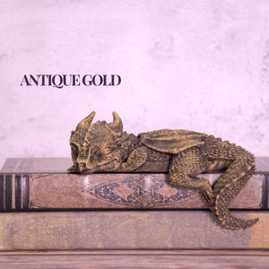 May include: An antique gold dragon figurine resting on two stacked, aged books. The dragon has detailed scales and wings, with the text "ANTIQUE GOLD" above. The books have a worn, vintage appearance.