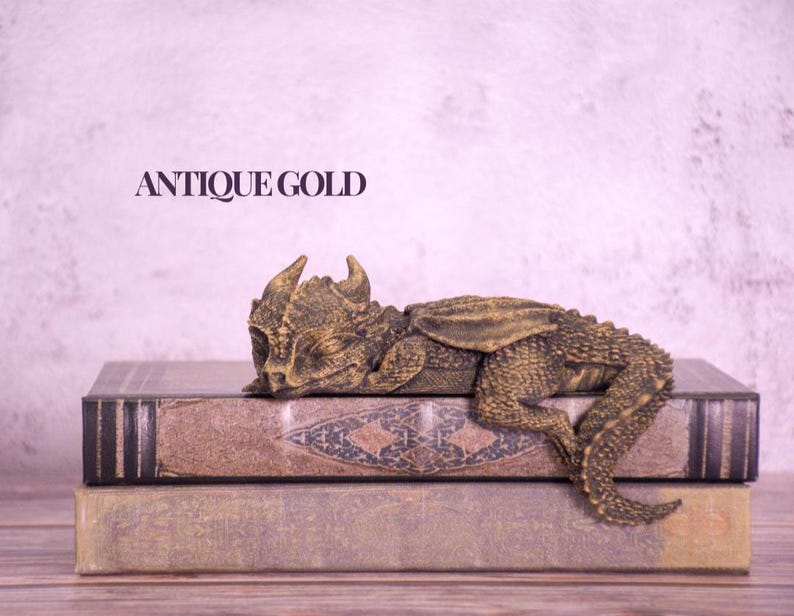 May include: An antique gold dragon figurine resting on two stacked, aged books. The dragon has detailed scales and wings, with the text "ANTIQUE GOLD" above. The books have a worn, vintage appearance.