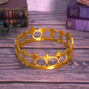 K&ouml;nnte beinhalten: Eine goldfarbene Krone mit kreisf&ouml;rmigem Design, mit offenen Kreisen und dekorativen Elementen. Die Krone liegt auf einer Holzoberfl&auml;che, mit B&uuml;chern im Hintergrund.