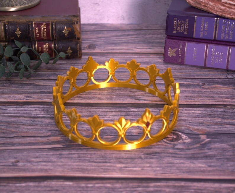 K&ouml;nnte beinhalten: Eine goldfarbene Krone mit kreisf&ouml;rmigem Design, mit offenen Kreisen und dekorativen Elementen. Die Krone liegt auf einer Holzoberfl&auml;che, mit B&uuml;chern im Hintergrund.