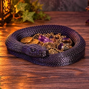 May include: A dark purple, coiled snake-shaped trinket dish with a textured, scale-like surface. The center of the dish holds a collection of gold-toned coins, a gold chain, a ring with a purple gemstone, and a raw purple amethyst crystal.