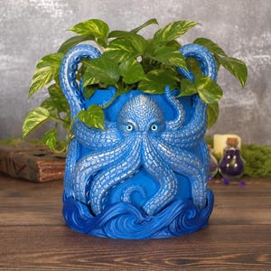 May include: A cylindrical blue planter featuring a sculpted octopus with silver-toned suction cups and bright blue eyes. The base of the planter is molded to resemble stylized ocean waves, and it contains a green and yellow variegated pothos plant.