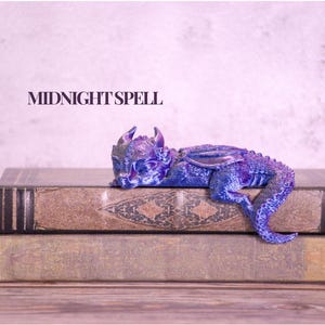 May include: A detailed, blue and purple dragon figurine rests on top of two antique books. The dragon has intricate scales and wings. The text "MIDNIGHT SPELL" is displayed above the books. The books have a worn, vintage appearance.