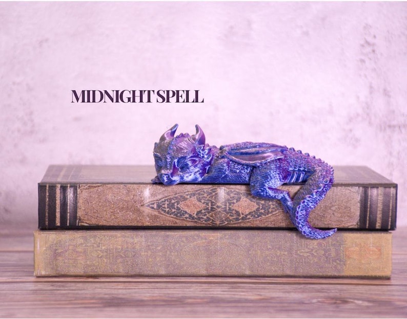 May include: A detailed, blue and purple dragon figurine rests on top of two antique books. The dragon has intricate scales and wings. The text "MIDNIGHT SPELL" is displayed above the books. The books have a worn, vintage appearance.