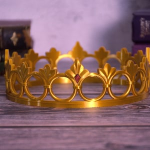 K&ouml;nnte beinhalten: Eine goldfarbene Krone mit B&ouml;gen und Fleur-de-Lis-Motiven. Ein kleiner roter Edelstein befindet sich in der Mitte der Vorderseite. Die Krone liegt auf einer Holzoberfl&auml;che.