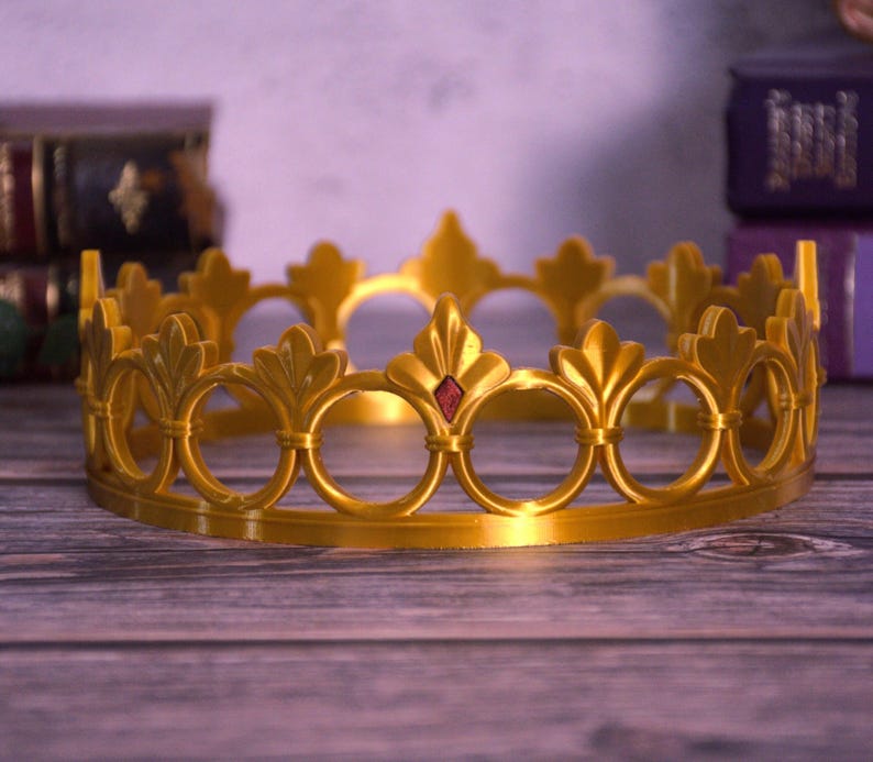K&ouml;nnte beinhalten: Eine goldfarbene Krone mit B&ouml;gen und Fleur-de-Lis-Motiven. Ein kleiner roter Edelstein befindet sich in der Mitte der Vorderseite. Die Krone liegt auf einer Holzoberfl&auml;che.
