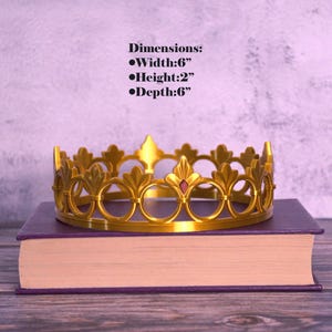 K&ouml;nnte beinhalten: Eine goldfarbene Krone mit dekorativem Design, die auf einem lila Buch liegt. Die Krone ist 15 cm breit, 5 cm hoch und 15 cm tief. Sie hat einen roten Edelstein in der Mitte. Abmessungen: Breite:6", H&ouml;he:2", Tiefe:6".
