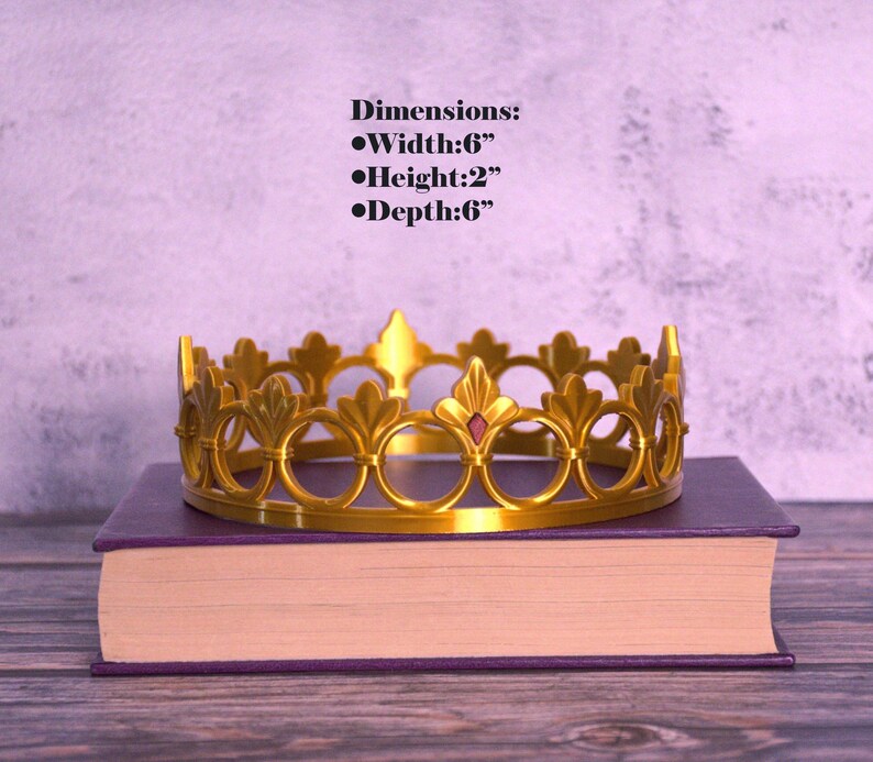 K&ouml;nnte beinhalten: Eine goldfarbene Krone mit dekorativem Design, die auf einem lila Buch liegt. Die Krone ist 15 cm breit, 5 cm hoch und 15 cm tief. Sie hat einen roten Edelstein in der Mitte. Abmessungen: Breite:6", H&ouml;he:2", Tiefe:6".
