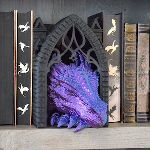 May include: A black gothic arch bookend featuring a detailed, iridescent purple dragon head emerging from the center. The dragon has textured scales and sharp claws visible at the base. It is positioned between two black books with dragon silhouettes.