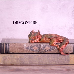 May include: A small, detailed dragon figurine in shades of red and green rests atop two antique-looking books. The figurine is lying down, with its head resting on the top book. The text "DRAGON FIRE" is displayed above the books.