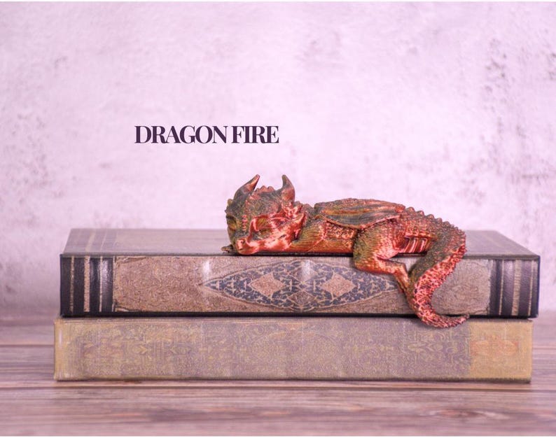 May include: A small, detailed dragon figurine in shades of red and green rests atop two antique-looking books. The figurine is lying down, with its head resting on the top book. The text "DRAGON FIRE" is displayed above the books.