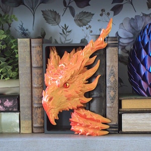 May include: A decorative bookend featuring a sculpted dragon head with vibrant orange and yellow flame-like textures, a single piercing red eye, and a sharp beak, positioned between two vintage-style books on a shelf.