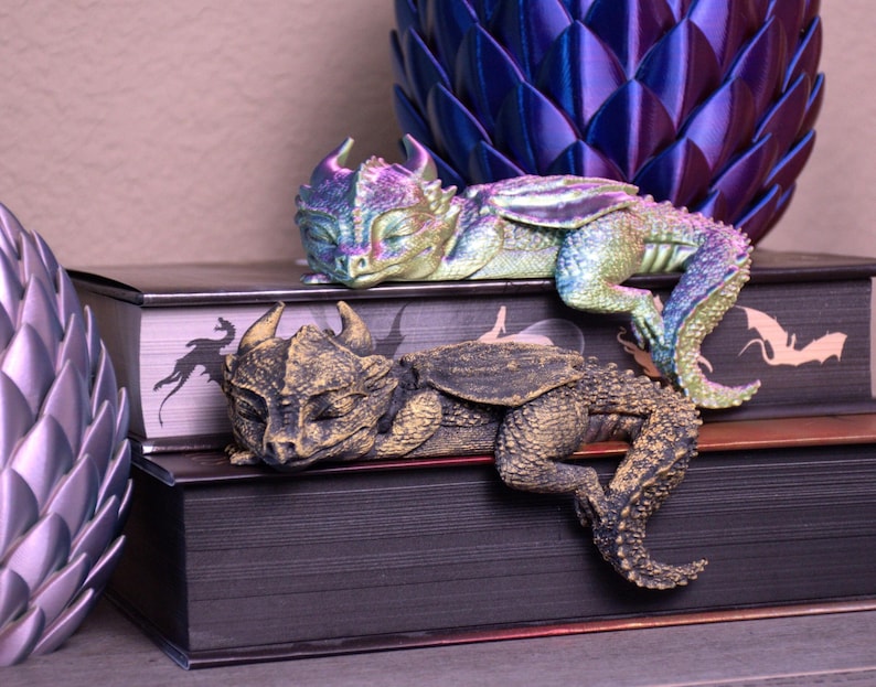 May include: Two decorative dragon figurines rest on stacked books. One dragon is gold and bronze, the other is iridescent with shades of green, blue, and purple. The books have a dark cover with a dragon silhouette.
