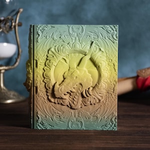 May include: A fantasy-themed book with a dragon design. The cover features a detailed relief of a dragon's head within an ornate frame, with a gradient of colors from green to yellow to brown. The book is on a wooden surface.