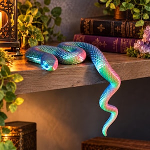 May include: A flexible, articulated snake figurine featuring a vibrant, iridescent gradient finish that shifts between shades of blue, green, and pink. The surface is covered in a detailed, repeating scale pattern, and the body is coiled.