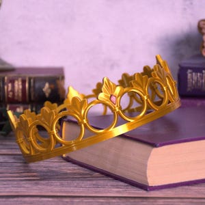 K&ouml;nnte beinhalten: Eine goldfarbene Krone mit kreisf&ouml;rmigen und blattf&ouml;rmigen Designs, akzentuiert mit einem kleinen rosa Edelstein. Die Krone liegt auf einem lila Buch, was auf ein Thema von K&ouml;nigtum oder Fantasie hindeutet. Im Hintergrund sind weitere B&uuml;cher und dekorative Elemente zu sehen.