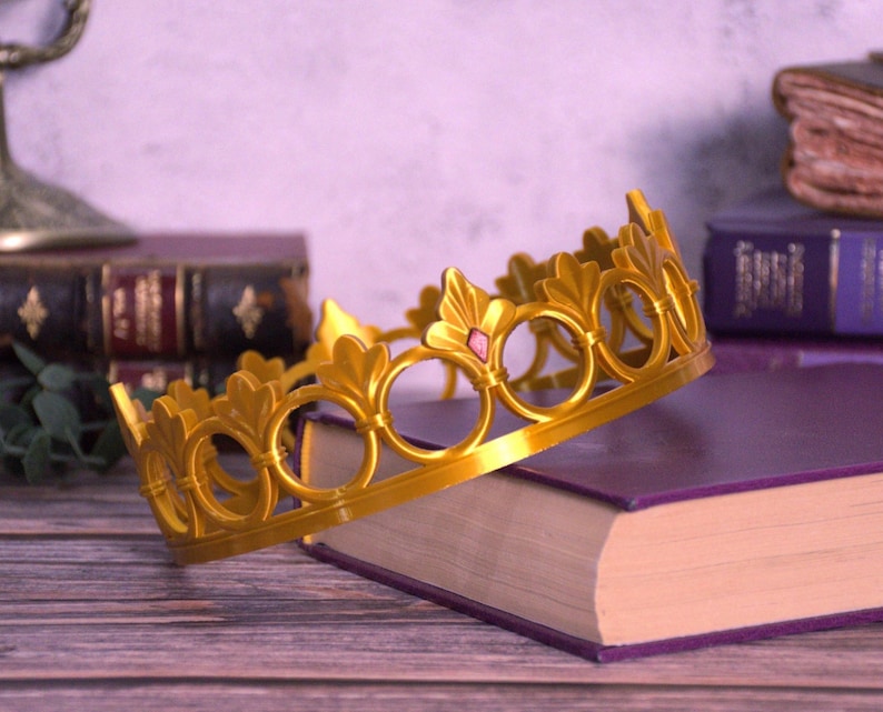 K&ouml;nnte beinhalten: Eine goldfarbene Krone mit kreisf&ouml;rmigen und blattf&ouml;rmigen Designs, akzentuiert mit einem kleinen rosa Edelstein. Die Krone liegt auf einem lila Buch, was auf ein Thema von K&ouml;nigtum oder Fantasie hindeutet. Im Hintergrund sind weitere B&uuml;cher und dekorative Elemente zu sehen.