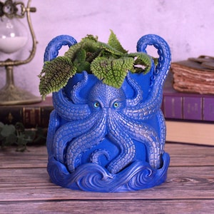 May include: A blue octopus-shaped planter with green leafy plants. The planter has a detailed design of an octopus with blue eyes and tentacles. The base of the planter features a wave design. The planter is made of a durable material.