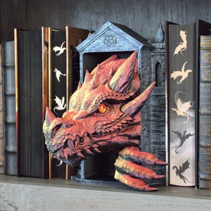 May include: A red dragon head sculpture emerging from a dark gray stone-like archway bookend. The dragon features textured scales, sharp horns, and a single glowing orange eye. The piece is positioned between two black books with dragon silhouettes.