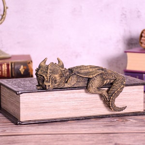 May include: A detailed, bronze-colored dragon figurine resting on a large, antique book. The dragon has textured scales and horns. The book has a decorative cover and aged pages. Other books and a candle holder are in the background.